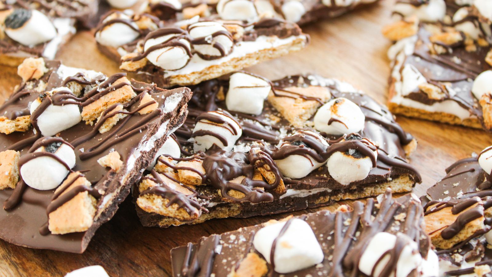 Pieces of chocolate bark topped with mini marshmallows, graham cracker pieces, and drizzled chocolate arranged on a wooden surface.
