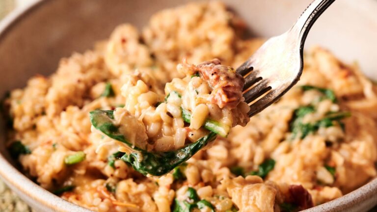 A close-up of a fork holding a bite of creamy risotto with spinach and pieces of sun-dried tomato, above a bowl filled with the same dish.