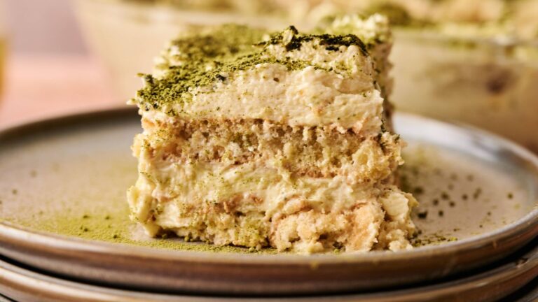 A slice of matcha tiramisu sits on a plate, showing layers of creamy filling and cake, with green matcha powder sprinkled on top.