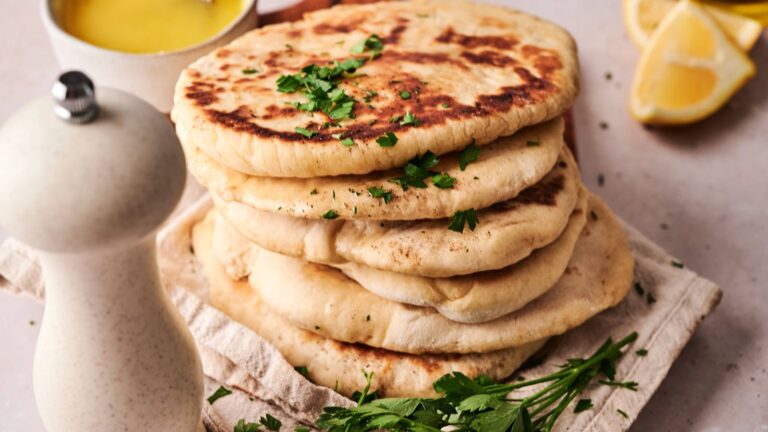 A stack of flatbreads garnished with chopped parsley sits on a cloth, with a pepper grinder, parsley, lemon wedges, and a bowl of yellow sauce nearby.