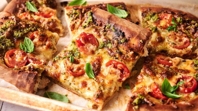 Rectangular pizza slices topped with melted cheese, cherry tomatoes, pesto sauce, and fresh basil leaves on a baking tray.
