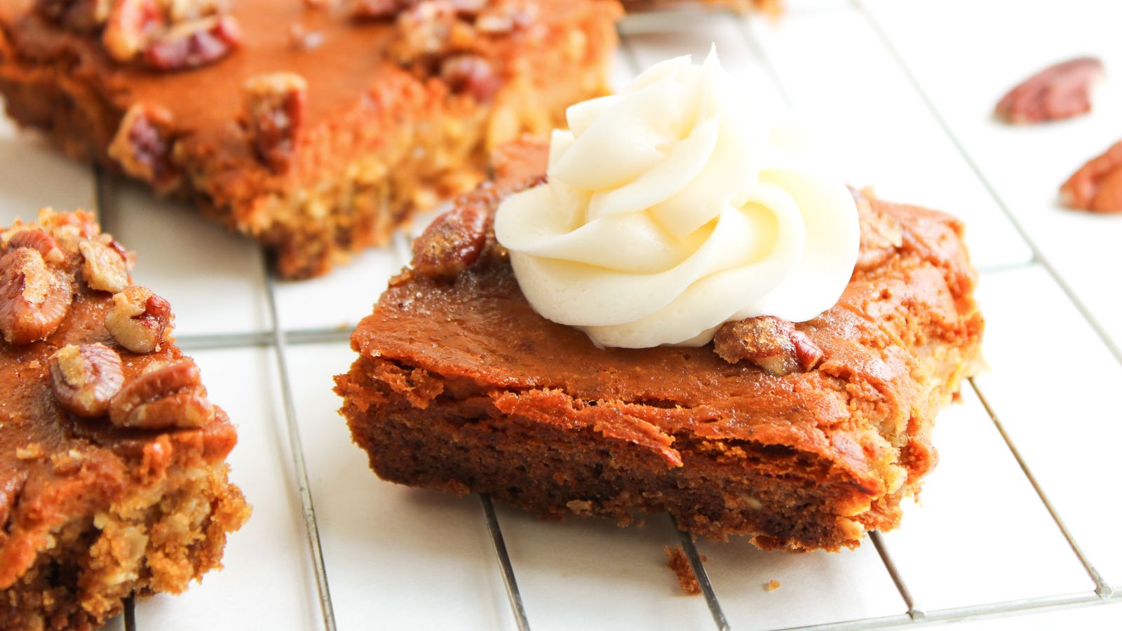 A square piece of pumpkin dessert bar topped with a swirl of white frosting and pecans sits on a cooling rack.