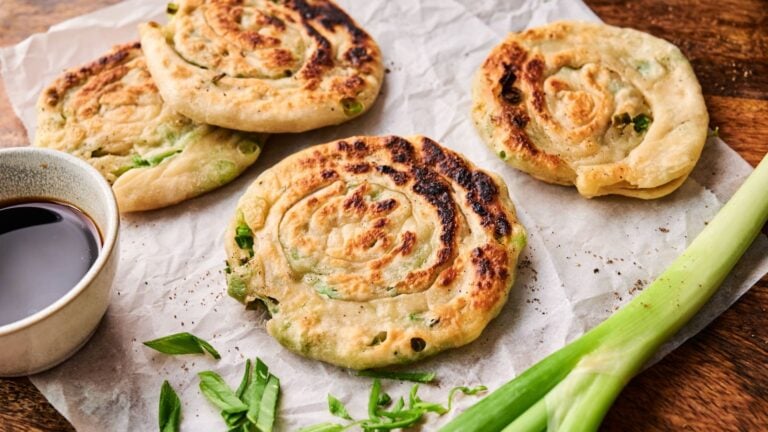 Four scallion pancakes on parchment paper with a small bowl of dipping sauce and fresh green onions beside them.