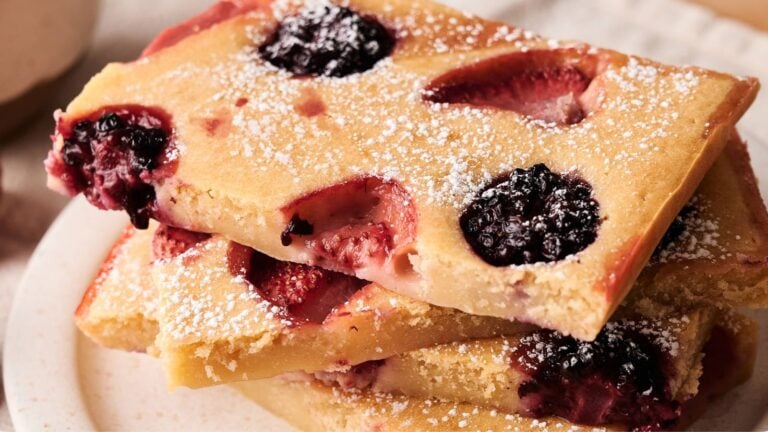 Three rectangular pieces of fruit-studded cake, topped with powdered sugar, are stacked on a white plate. Each piece has visible blackberries and strawberries.