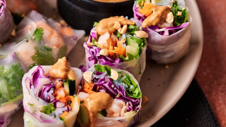 Fresh spring rolls filled with shrimp, purple cabbage, carrots, lettuce, and cucumbers, topped with peanut sauce and crushed peanuts, served on a plate with dipping sauce.