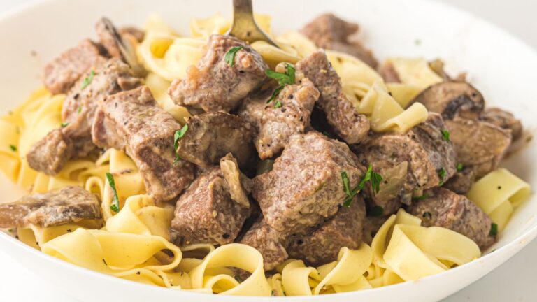 A close-up of beef stroganoff with wide egg noodles, featuring chunks of beef and mushrooms in a creamy sauce, garnished with herbs.