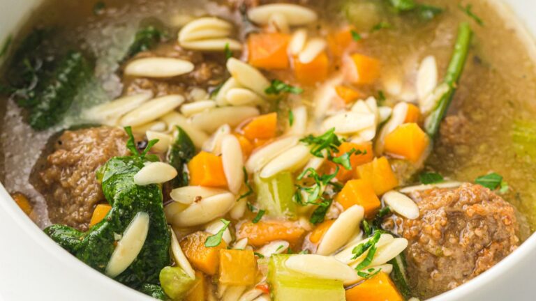 A close-up of a bowl of soup with meatballs, sliced almonds, chopped carrots, celery, greens, and broth.