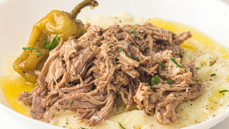 Shredded pot roast served over mashed potatoes with a whole pepper and garnished with chopped herbs, in a white bowl.
