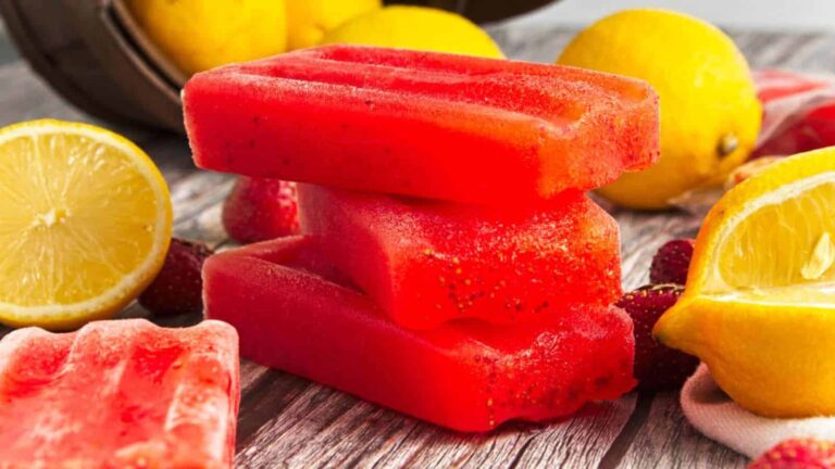 Three red popsicles are stacked on a wooden surface surrounded by halved and whole lemons and strawberries.