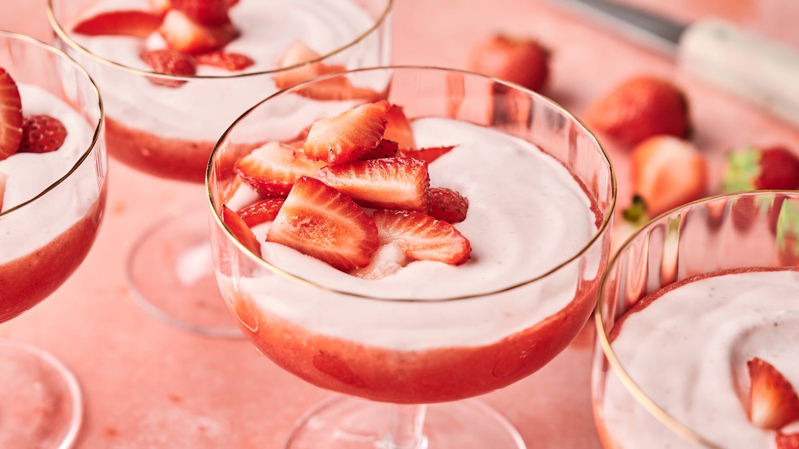 Three glass dessert cups filled with strawberry mousse, topped with fresh strawberry slices on a pink surface.