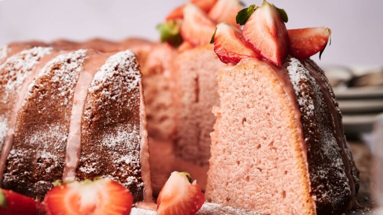 A strawberry bundt cake dusted with powdered sugar, topped with sliced strawberries, with one slice cut and partially removed.