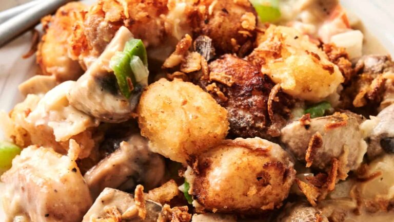 Close-up of a casserole dish containing tater tots, pieces of meat, mushrooms, green peppers, and a creamy sauce, topped with crispy fried onions.