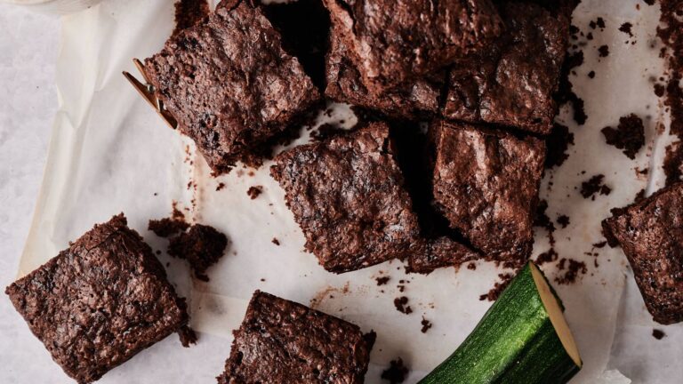 Several chocolate brownies are arranged on parchment paper next to a piece of fresh zucchini.