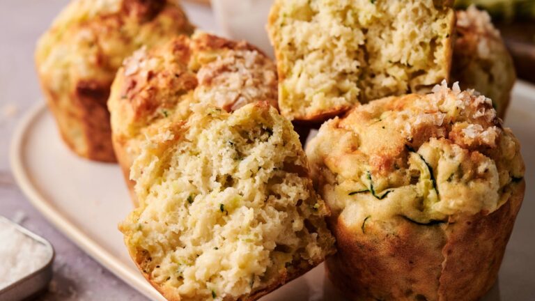 A close-up of several zucchini muffins, some whole and some halved, showing their moist, textured interior on a white plate.