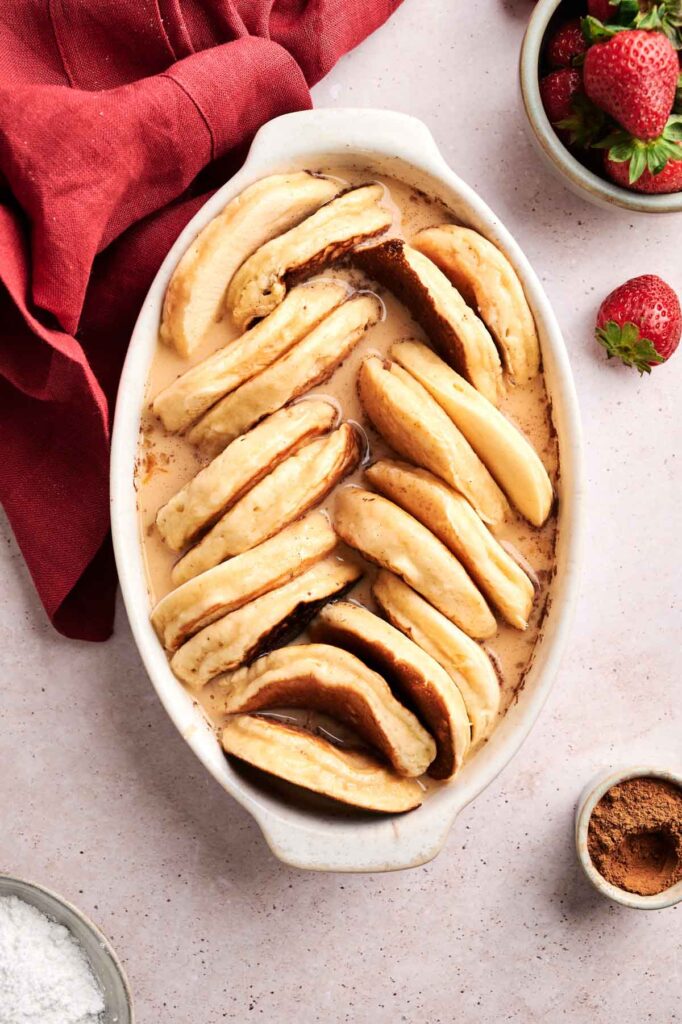 A white oval baking dish filled with sliced baked bananas arranged upright, perfect for a pancake casserole, is surrounded by bowls of strawberries, cinnamon, and a red cloth napkin.