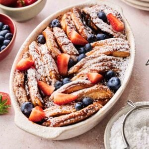 A baked pancake casserole of sliced French toast topped with powdered sugar, fresh strawberries, and blueberries, served with bowls of fruit and utensils nearby.