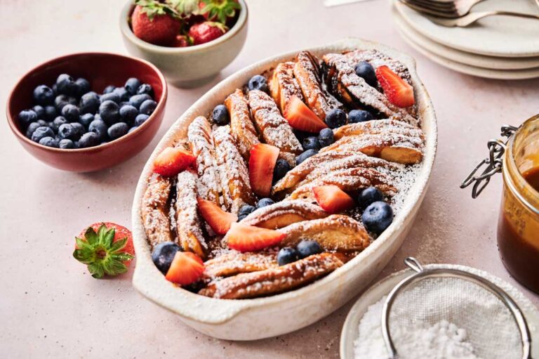 An oval baking dish holds a pancake casserole with baked pastries dusted in powdered sugar, topped with sliced strawberries and blueberries; bowls of fresh fruit and a jar of sauce sit nearby.