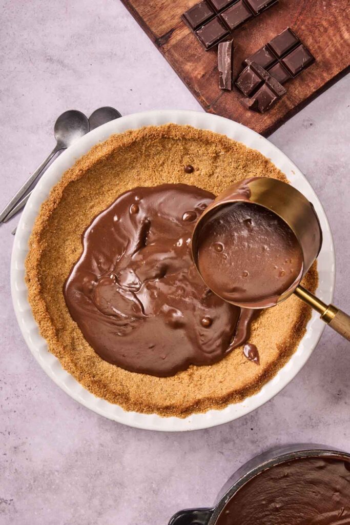 A graham cracker crust in a pie dish is being filled with rich chocolate filling from a measuring cup, creating the base for a delicious Chocolate Cream Pie, with chocolate squares on a cutting board nearby.