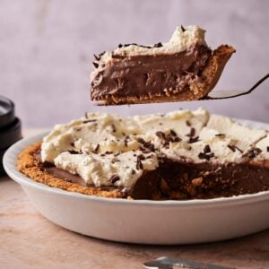 A slice of Chocolate Cream Pie with whipped topping is being lifted from a pie dish, surrounded by scattered chocolate pieces on the table.