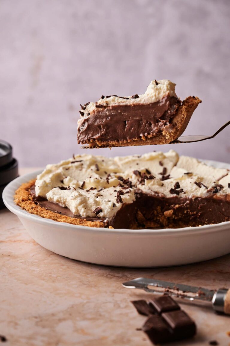 A slice of Chocolate Cream Pie with whipped topping is being lifted from a pie dish, surrounded by scattered chocolate pieces on the table.