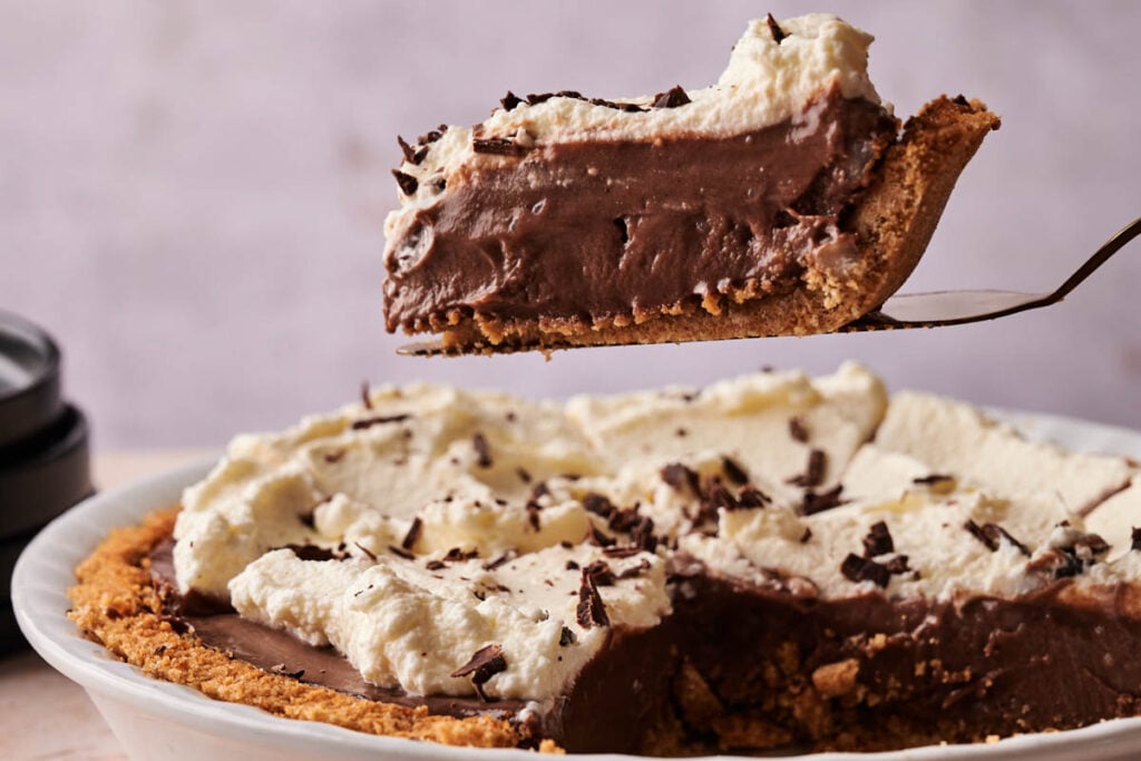 A slice of Chocolate Cream Pie with whipped cream and chocolate shavings is being lifted from a pie dish.