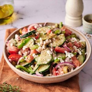A bowl of Greek Salad with cucumbers, tomatoes, red onions, olives, crumbled feta cheese, and herbs sits on a brown napkin with a pepper grinder and olive oil in the background.