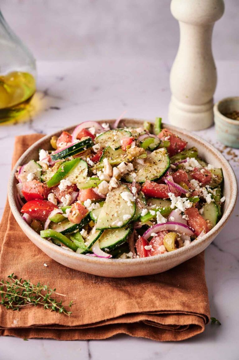 A bowl of Greek Salad with cucumbers, tomatoes, red onions, olives, crumbled feta cheese, and herbs sits on a brown napkin with a pepper grinder and olive oil in the background.