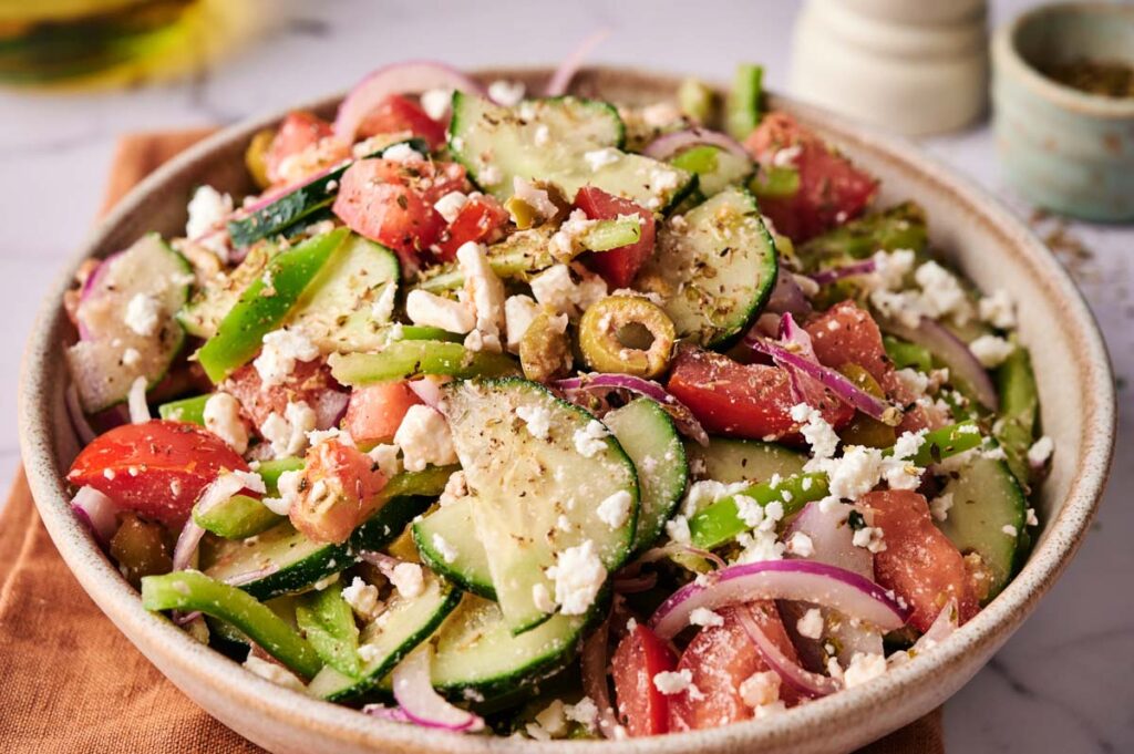 A bowl of Greek Salad with sliced cucumbers, tomatoes, red onions, green bell peppers, olives, crumbled feta cheese, and seasoning.
