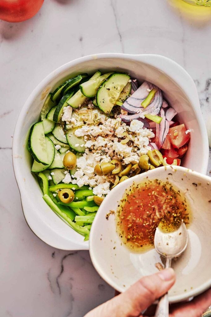 A bowl of Greek Salad with cucumbers, tomatoes, red onions, green bell peppers, olives, and feta cheese sits beside a hand holding a bowl of vinaigrette dressing.
