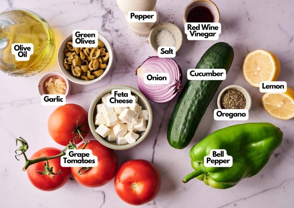 A flat lay of Greek Salad ingredients, including olive oil, green olives, garlic, grape tomatoes, feta cheese, onion, cucumber, bell pepper, salt, pepper, oregano, lemon, and red wine vinegar.