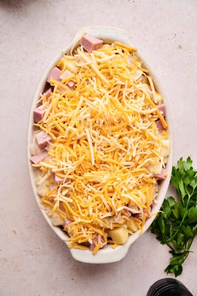 Oval baking dish filled with cubed ham, pasta, and shredded cheese, ready to be baked; reminiscent of a classic Ham and Potato Casserole. A bunch of fresh parsley sits nearby on the countertop.