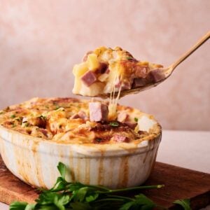 A serving spoon lifts a portion of cheesy Ham and Potato Casserole with diced ham and potatoes from a white dish, placed on a wooden board with fresh parsley nearby.