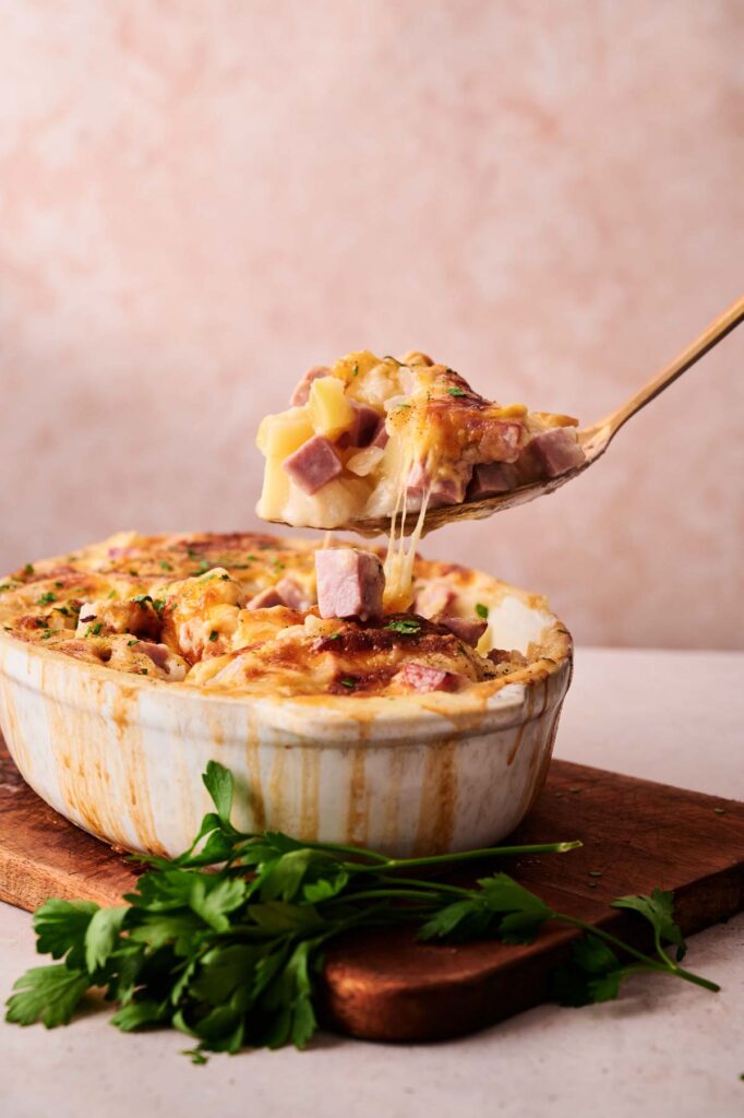 A serving spoon lifts a portion of cheesy Ham and Potato Casserole with diced ham and potatoes from a white dish, placed on a wooden board with fresh parsley nearby.