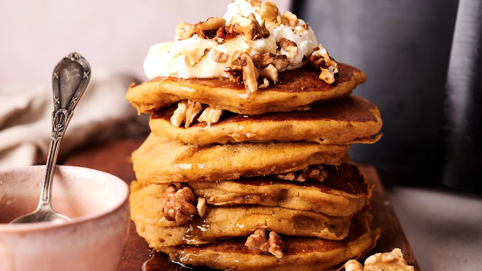 A stack of pancakes topped with whipped cream, walnuts, and syrup sits on a wooden board next to a pink bowl and a spoon.
