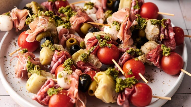 A plate of antipasto skewers with cherry tomatoes, artichoke hearts, olives, mozzarella balls, cured meats, and pesto sauce.