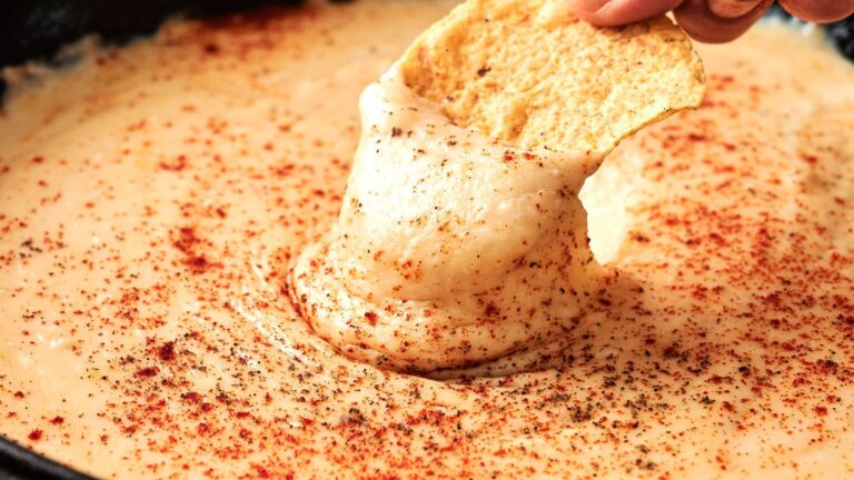 A hand dips a tortilla chip into a creamy, melted cheese dip topped with a sprinkle of red and black spices.