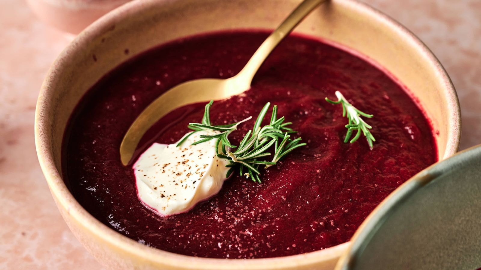 A bowl of beet soup is garnished with a dollop of cream, black pepper, and fresh rosemary, with a gold spoon resting inside the bowl.