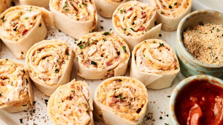 A plate of tortilla pinwheels filled with a creamy mixture, garnished with herbs, next to a bowl of breadcrumbs and a bowl of red sauce.