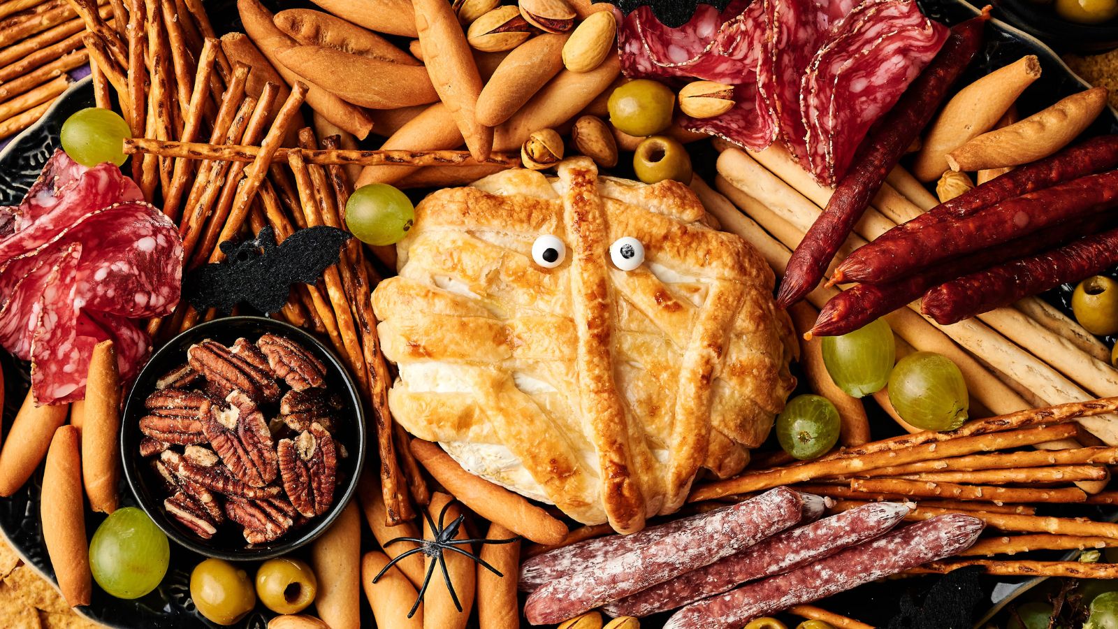 A cheese wheel wrapped in pastry to look like a mummy with candy eyes, surrounded by cured meats, breadsticks, pretzels, grapes, pecans, olives, and Halloween decorations.