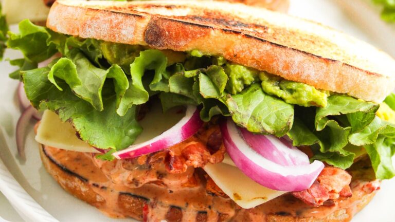 Close-up of a sandwich with toasted bread, leafy lettuce, sliced red onion, cheese, bacon, guacamole, and creamy spread.