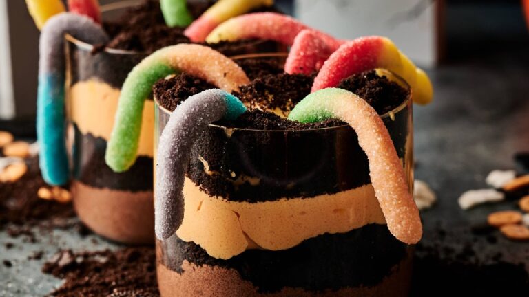 Glass cups filled with layered chocolate and cream dessert, topped with crushed cookies and decorated with colorful gummy worms.