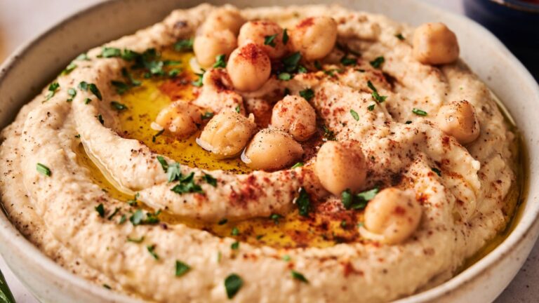 A bowl of hummus topped with whole chickpeas, olive oil, chopped parsley, and sprinkled with paprika.