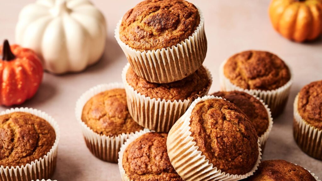 A stack of pumpkin muffins in paper liners is displayed on a light surface, with small decorative pumpkins in the background.