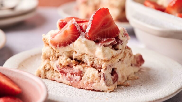 A slice of strawberry tiramisu sits on a white plate, topped with fresh strawberry halves and dusted with powdered sugar.
