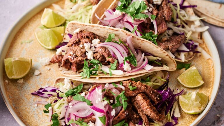 Two beef tacos on a plate topped with shredded cabbage, sliced red onions, crumbled cheese, and fresh cilantro, served with lime wedges.