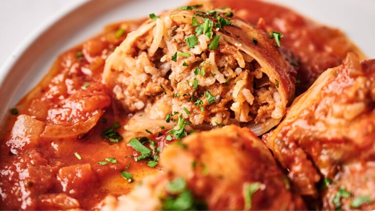 A close-up of stuffed cabbage rolls filled with rice and ground meat, topped with tomato sauce and chopped parsley on a white plate.