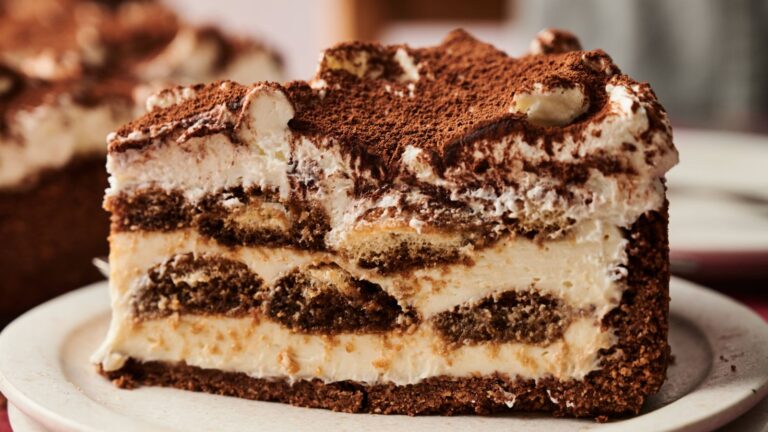 A close-up of a slice of tiramisu cheesecake on a white plate, showing layers of creamy filling, soaked cake, and a dusting of cocoa powder on top.