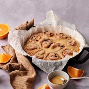 A round pan of glazed Orange Rolls lined with parchment paper sits on a marble surface, surrounded by orange slices, a small bowl of glaze, and a brown napkin.