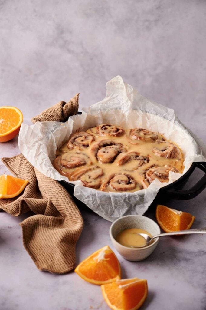 A round pan of glazed Orange Rolls lined with parchment paper sits on a marble surface, surrounded by orange slices, a small bowl of glaze, and a brown napkin.