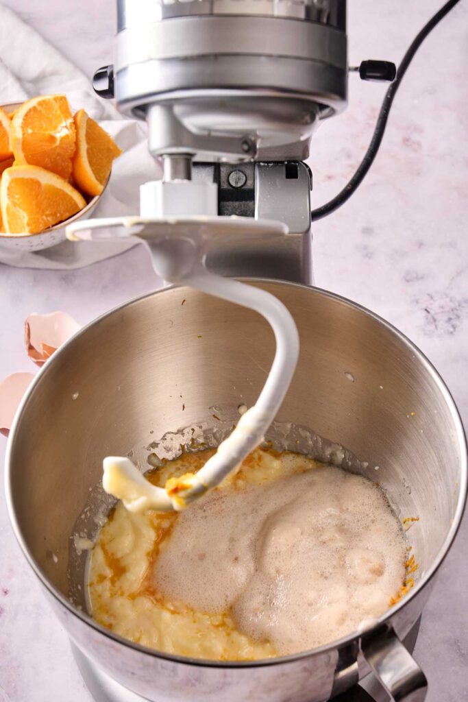 A stand mixer with paddle attachment blending ingredients for Orange Rolls; orange slices and eggshells are visible nearby on the counter.